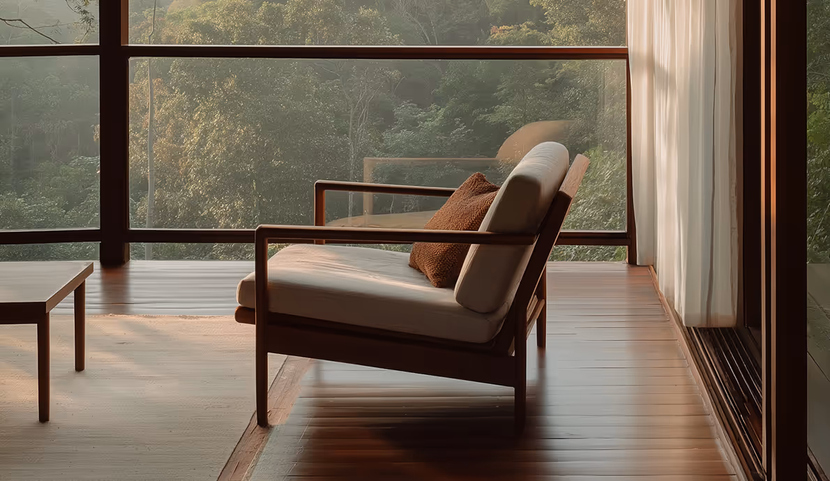 Cozy wooden armchair with beige cushions and a brown pillow near large glass windows overlooking a green forest.