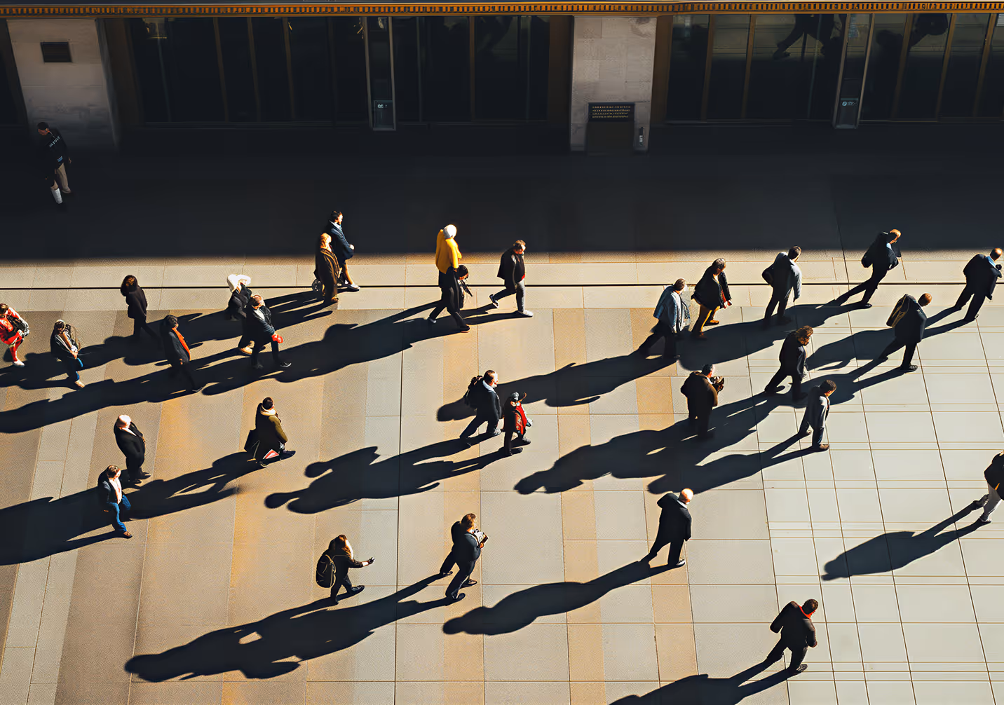 Overhead view of people walking on a tiled pavement casting long shadows in bright sunlight.